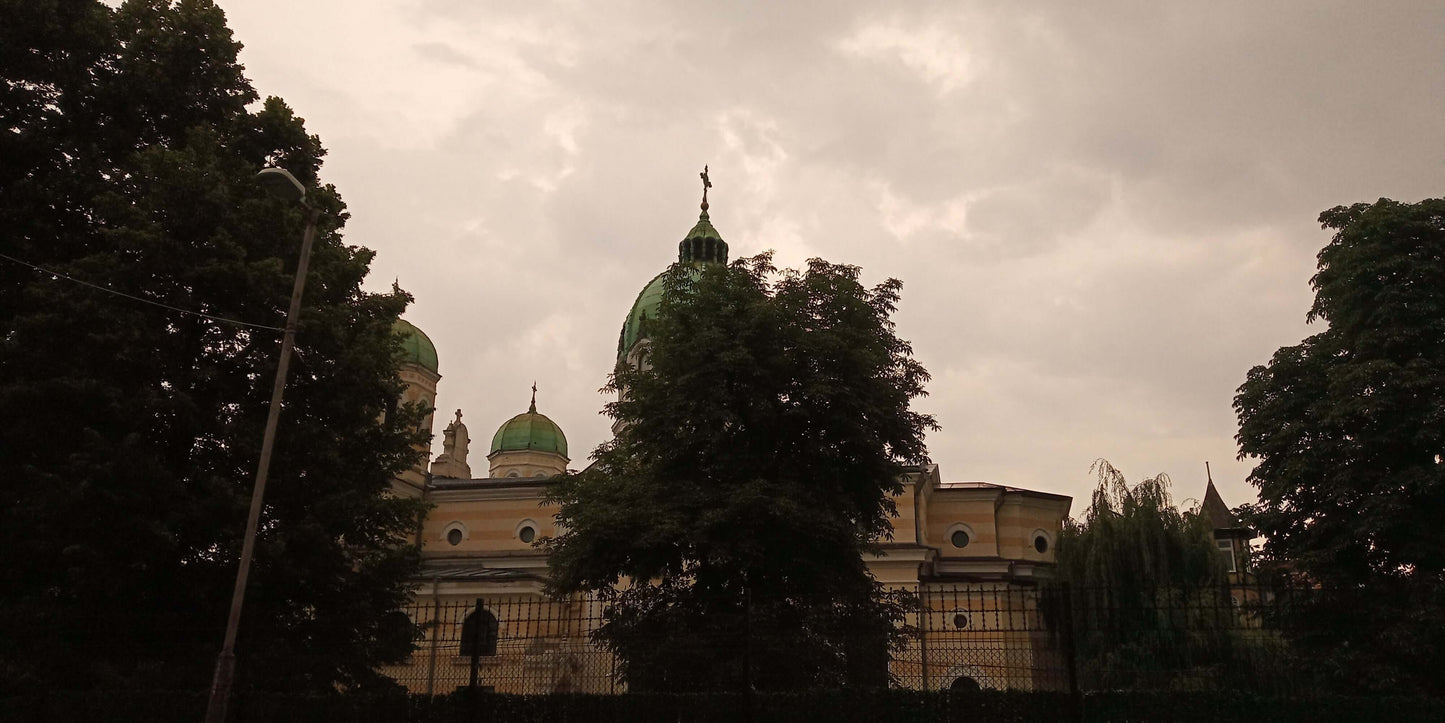 Stormy sky over a historic building with green domes, surrounded by lush trees.